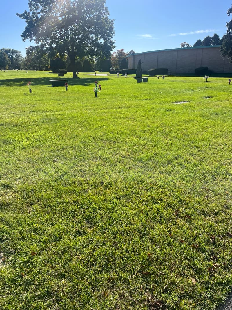 A wide lawn with flat markers and flower vases, a large shade tree and a long mausoleum building beyond.