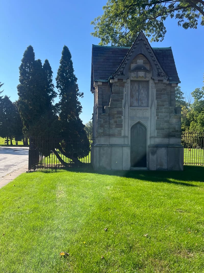 A small stone mausoleum with a steep slate roof, beside tall evergreens.